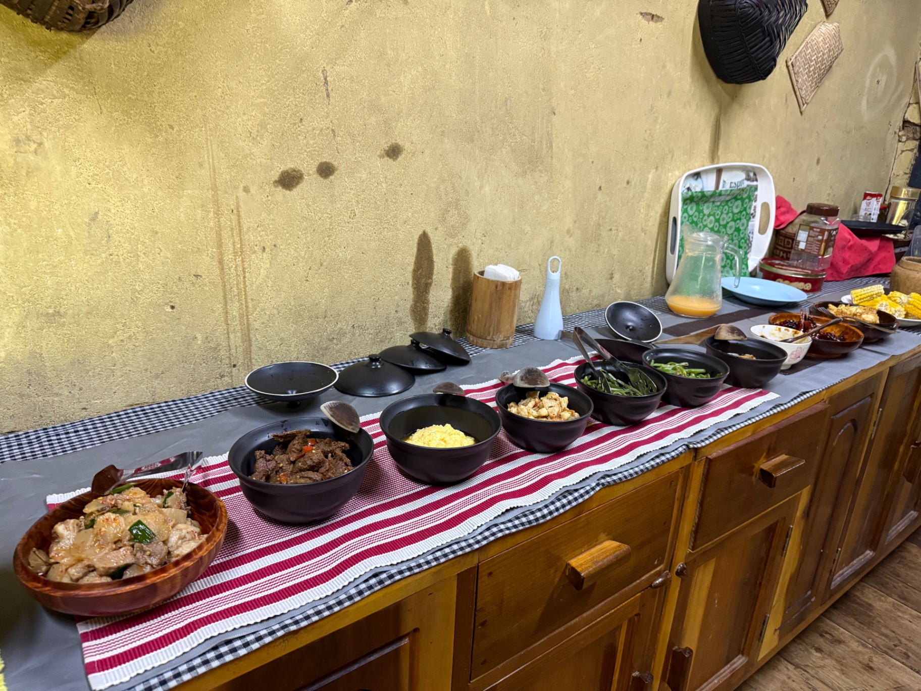 A row of dishes laid out on a wooden counter: meat, beef with chilies, rice, cheese dishes and green beans