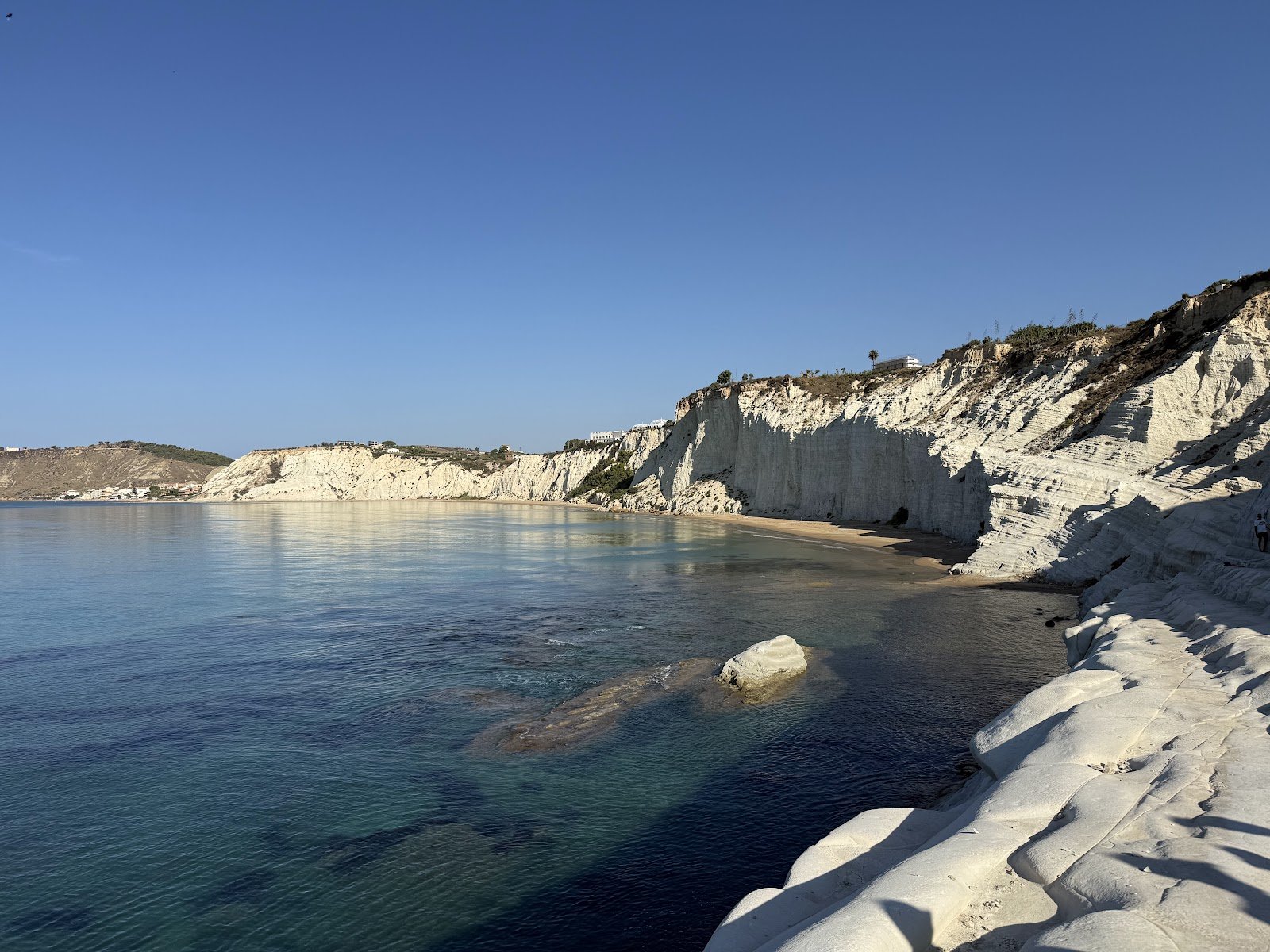 The layered white cliffs and rock steps of Scala dei Turchi curving around a clear turquoise bay under a deep blue sky